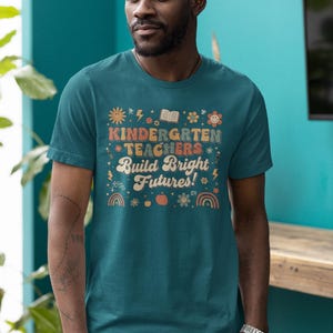 May include: Teal t-shirt with the text "KINDERGARTEN TEACHERS Build Bright Futures!" in a retro font. The design includes illustrations of a sun, book, flowers, and rainbows. The shirt is worn by a person.
