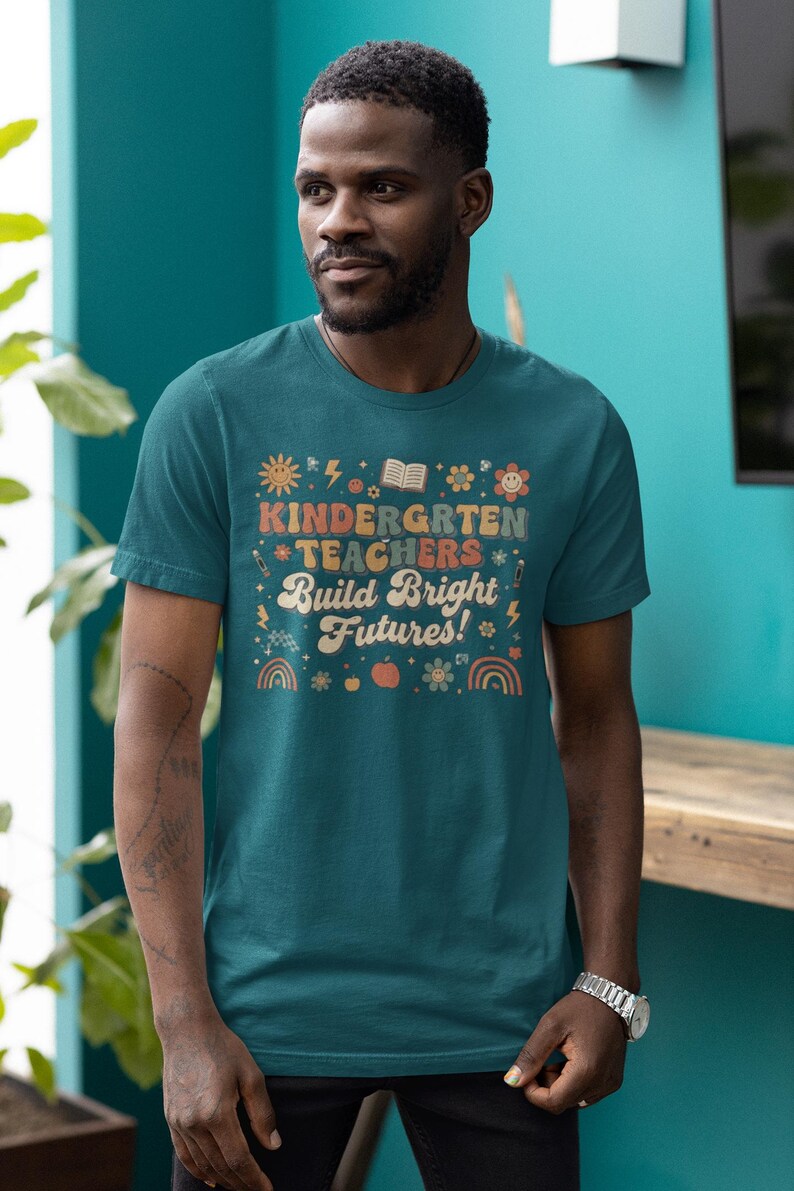 May include: Teal t-shirt with the text "KINDERGARTEN TEACHERS Build Bright Futures!" in a retro font. The design includes illustrations of a sun, book, flowers, and rainbows. The shirt is worn by a person.