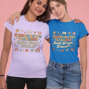 May include: Two women wearing matching t-shirts. One shirt is light purple, the other is blue. Both shirts have the text "KINDERGARTEN TEACHERS Build Bright Futures!" in a colorful, retro design. The women are smiling and embracing each other.