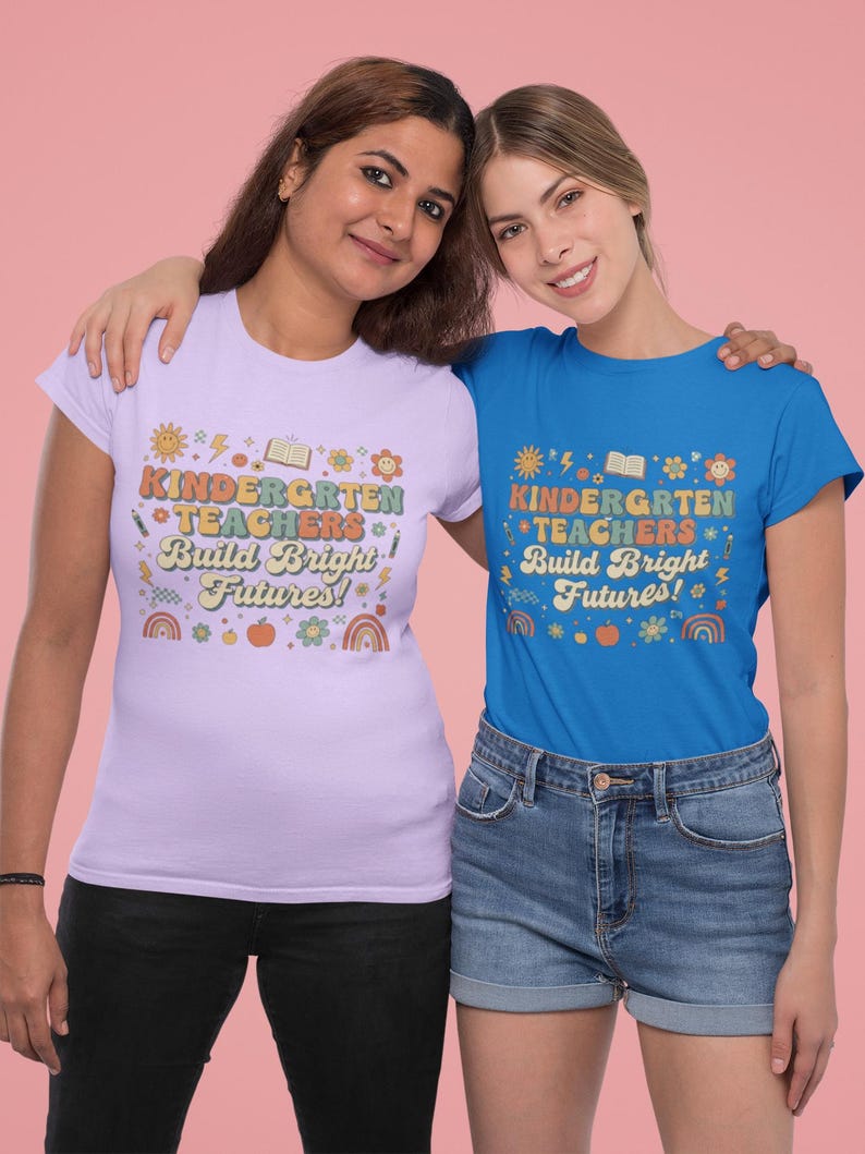 May include: Two women wearing matching t-shirts. One shirt is light purple, the other is blue. Both shirts have the text "KINDERGARTEN TEACHERS Build Bright Futures!" in a colorful, retro design. The women are smiling and embracing each other.