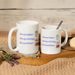 Può includere: Due tazze di ceramica bianca con le parole "Dream Maker, Always Giving, Devoted Heart" in testo rosso e blu. Ogni tazza ha un acchiappasogni e un cuore. Una tazza è più grande dell'altra e un cucchiaio è nella tazza più piccola.
