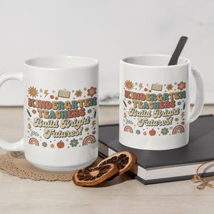 May include: Two white ceramic mugs with the text "KINDERGARTEN TEACHERS Build Bright Futures!" in a retro font. The mugs have a handle and are decorated with colorful illustrations of a sun, flowers, and rainbows. One mug has a spoon.