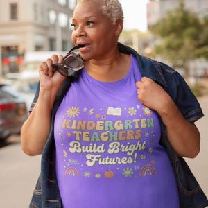 May include: A purple t-shirt with the text "KINDERGARTEN TEACHERS Build Bright Futures!" in colorful letters and various graphics. The shirt is worn with a denim jacket.