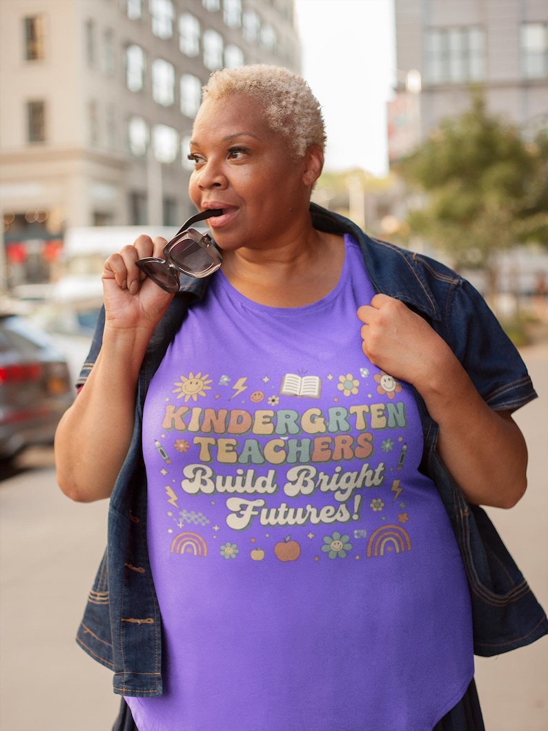 May include: A purple t-shirt with the text "KINDERGARTEN TEACHERS Build Bright Futures!" in colorful letters and various graphics. The shirt is worn with a denim jacket.