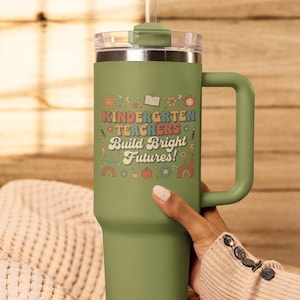 May include: A green insulated tumbler with a handle and a clear lid. The tumbler features the text "KINDERGARTEN TEACHERS Build Bright Futures!" in a colorful, retro font, along with various illustrations. A straw is inserted into the lid.