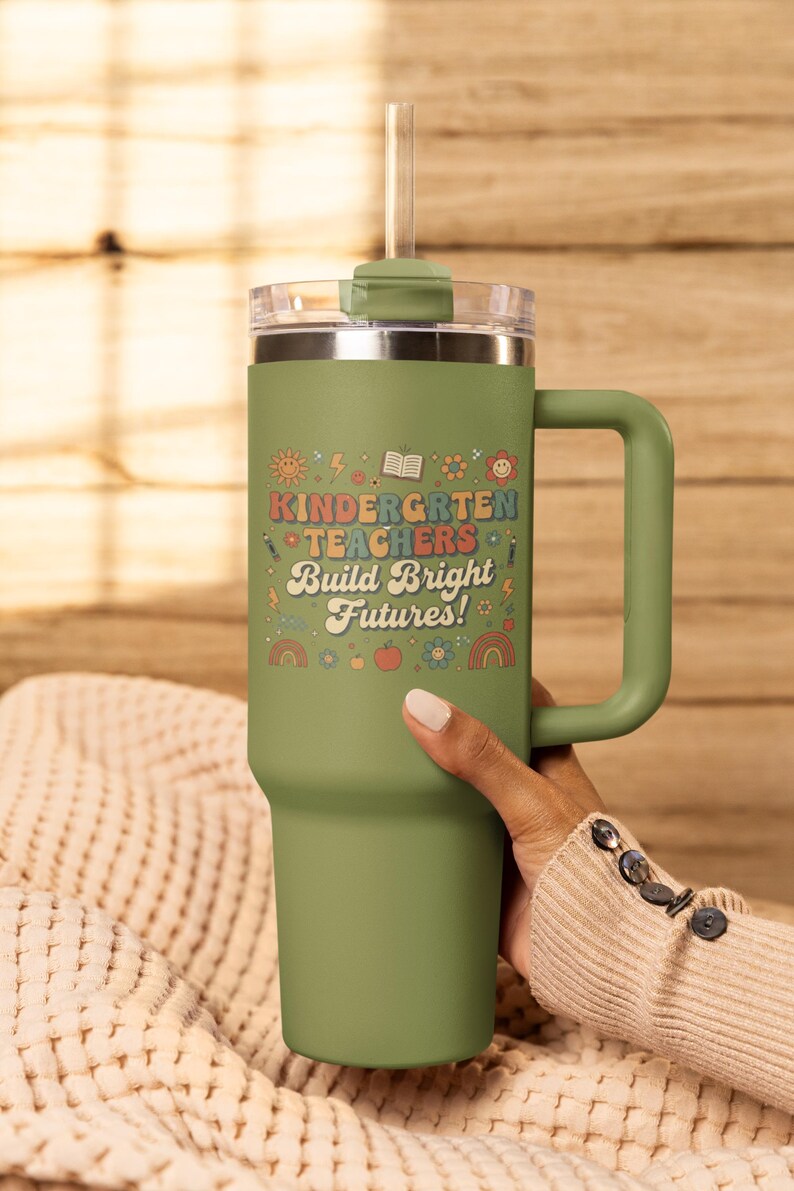 May include: A green insulated tumbler with a handle and a clear lid. The tumbler features the text "KINDERGARTEN TEACHERS Build Bright Futures!" in a colorful, retro font, along with various illustrations. A straw is inserted into the lid.