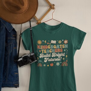 May include: A green t-shirt with the text "KINDERGARTEN TEACHERS Build Bright Futures!" in a colorful, retro design. The shirt is hanging on a hanger with a camera and a brown hat nearby. A denim jacket is also visible.