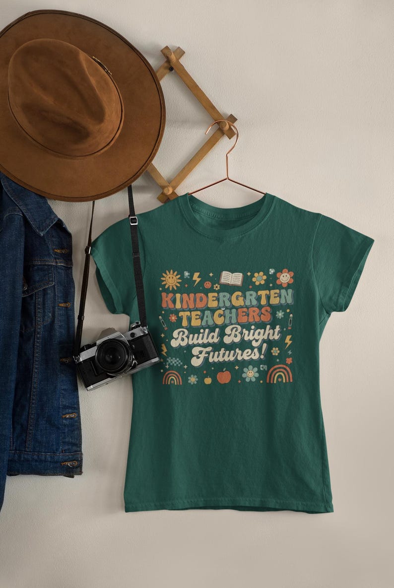 May include: A green t-shirt with the text "KINDERGARTEN TEACHERS Build Bright Futures!" in a colorful, retro design. The shirt is hanging on a hanger with a camera and a brown hat nearby. A denim jacket is also visible.
