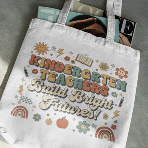 May include: White tote bag with the text "KINDERGARTEN TEACHERS Build Bright Futures!" in retro-style lettering. The design includes a smiling sun, flowers, rainbows, and an apple. The bag is filled with magazines.
