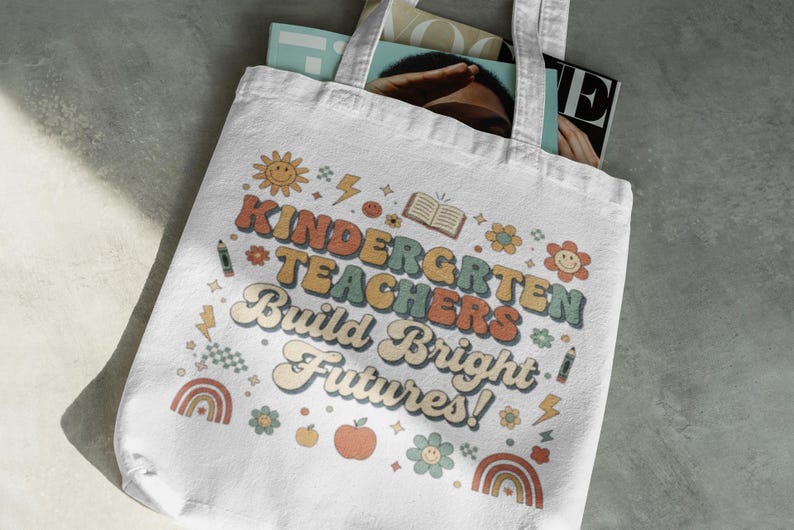 May include: White tote bag with the text "KINDERGARTEN TEACHERS Build Bright Futures!" in retro-style lettering. The design includes a smiling sun, flowers, rainbows, and an apple. The bag is filled with magazines.