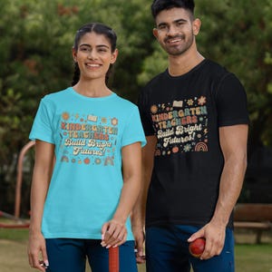 May include: Two people wearing matching t-shirts with the text "KINDERGARTEN TEACHERS Build Bright Futures!". One shirt is light blue, the other black. The design includes books, rainbows, and other colorful graphics. They are standing outside.