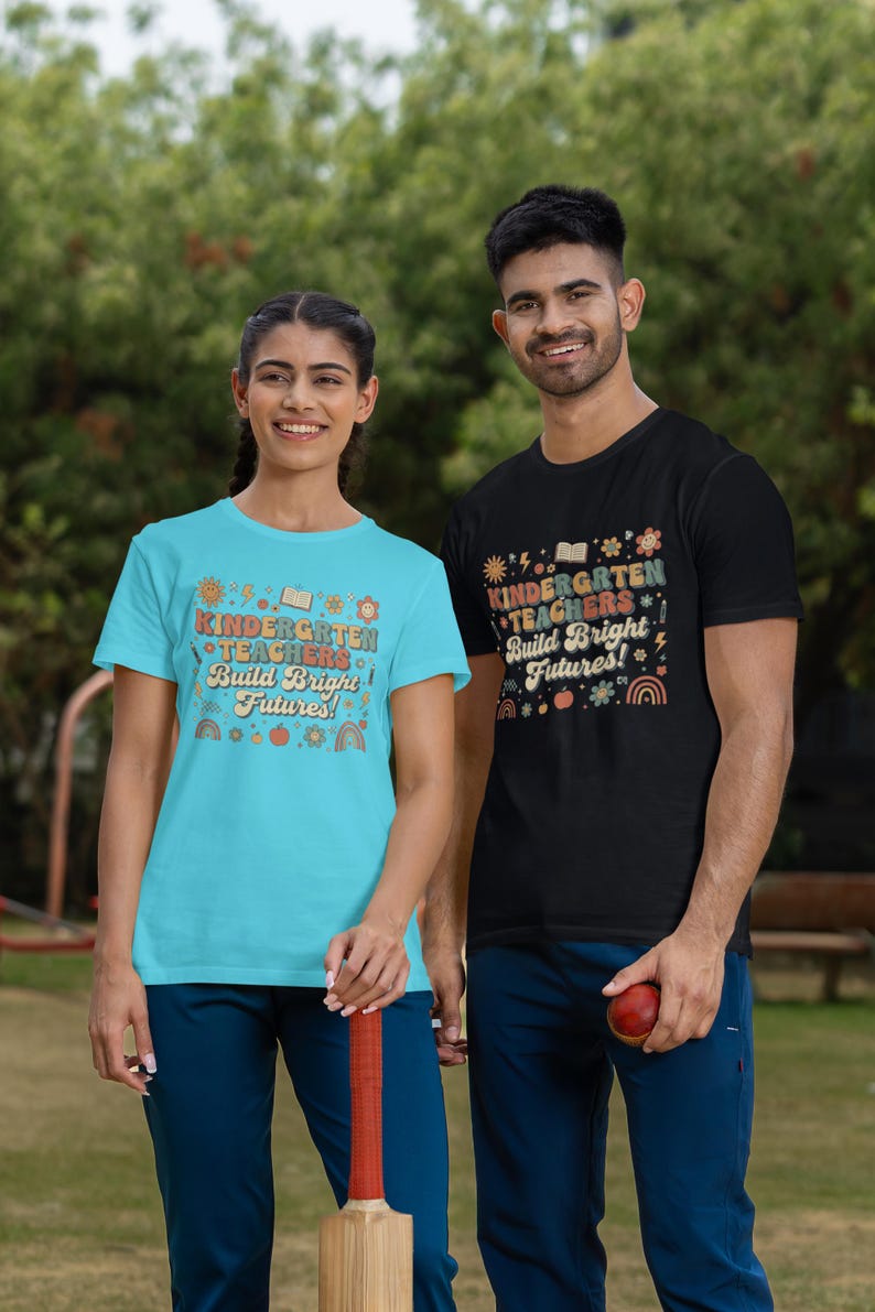 May include: Two people wearing matching t-shirts with the text "KINDERGARTEN TEACHERS Build Bright Futures!". One shirt is light blue, the other black. The design includes books, rainbows, and other colorful graphics. They are standing outside.