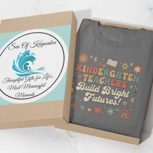 May include: A gray t-shirt with the text "KINDERGARTEN TEACHERS Build Bright Futures!" in colorful letters and graphics, inside a brown gift box with a blue and white label that reads "Sea Of Keepsakes".