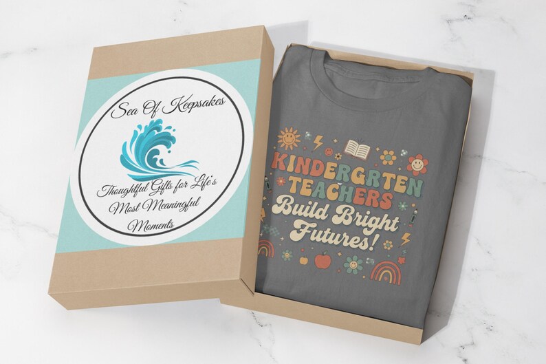 May include: A gray t-shirt with the text "KINDERGARTEN TEACHERS Build Bright Futures!" in colorful letters and graphics, inside a brown gift box with a blue and white label that reads "Sea Of Keepsakes".