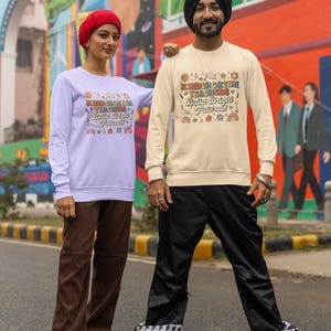 May include: Two people wearing matching sweatshirts with the text "Kindergarten Teachers Build Bright Futures!" in a colorful design. The sweatshirts are light purple and cream-colored. The person on the left is wearing brown pants and the person on the right is wearing black pants.
