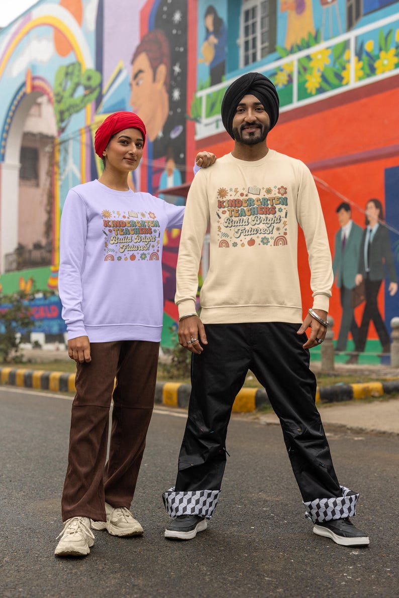 May include: Two people wearing matching sweatshirts with the text "Kindergarten Teachers Build Bright Futures!" in a colorful design. The sweatshirts are light purple and cream-colored. The person on the left is wearing brown pants and the person on the right is wearing black pants.
