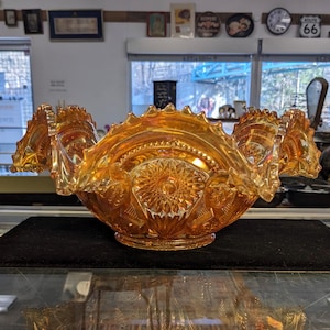 May include: An iridescent orange glass bowl with a ruffled edge. The bowl has a detailed pattern of floral and geometric designs. The bowl is sitting on a black surface.