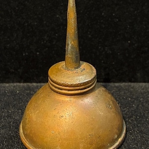 May include: A vintage, antique oil can with a brass finish. The can has a rounded base, a cylindrical body, and a long, tapered spout. The metal has a tarnished appearance, suggesting age and use. The oil can is likely used for lubricating machinery or tools.