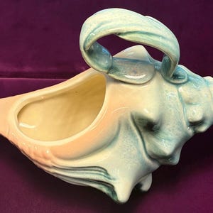 Op de afbeelding: Een keramische schelpvormige plantenbak met een handvat. De plantenbak is geschilderd in tinten perzik, crème en turquoise. De handgreep heeft een gebogen, boogvormige vorm. De plantenbak heeft een grote opening.