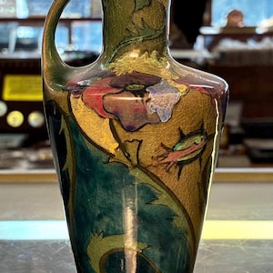 May include: An antique vase with a handle, featuring a floral design in shades of green, gold, and blue. The vase has a crackled texture and a glossy finish, showcasing a blend of artistry and vintage charm.