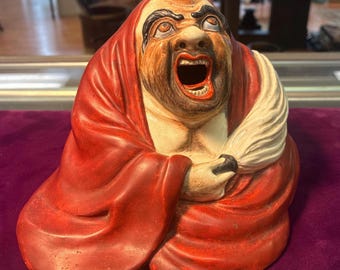 May include: A ceramic figurine of a seated figure with a wide open mouth, wearing a red robe and a white headdress. The figure has a detailed face with a beard and furrowed brow. The figurine is on a purple surface.