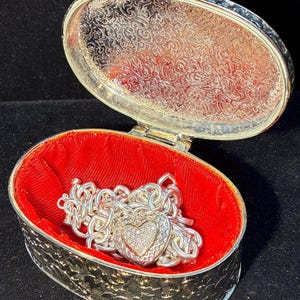 May include: An open, silver-colored oval jewelry box with a red velvet interior. A silver chain necklace with a heart-shaped pendant is inside. The pendant is encrusted with small, clear stones. The box has a decorative, engraved pattern.