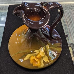 Puede incluir: Una jarra decorativa de cerámica marrón oscuro con un diseño floral amarillo y marrón. La jarra tiene un asa única y una base ancha y redonda. El diseño floral presenta un narciso amarillo con hojas verdes.