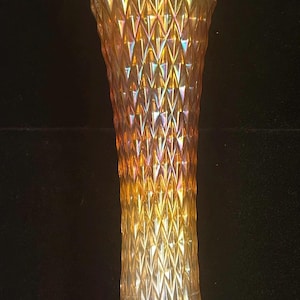 May include: A tall, iridescent glass vase with a diamond-patterned exterior. The vase has a flared top and a slender, elongated body. The colors shift from gold to orange and display a rainbow effect. The vase is likely used for decorative purposes.