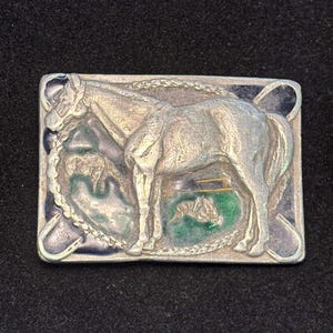 May include: Silver-toned rectangular belt buckle with a detailed relief of a horse. The horse is standing in a field with a rider. The buckle has a blue enamel border and a decorative chain-like edge. The horse is the focal point.