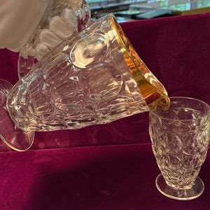 Puede incluir: Una jarra de cristal transparente con borde dorado vierte en un vaso a juego. Ambas piezas tienen un patrón de círculos repetidos. La jarra tiene un asa y una parte superior decorativa. El vaso está sobre un pie pequeño.