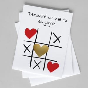 May include: White greeting cards with a tic-tac-toe design. The game board features black lines, red hearts, and a gold heart. The French text "Découvre ce que tu as gagné" is written at the top of the card.