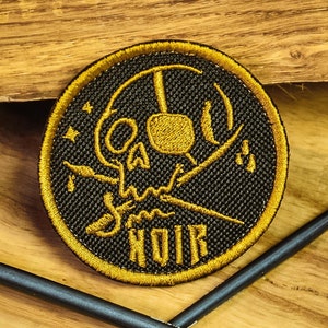 May include: A black and gold embroidered patch with a skull and crossbones design. The skull has a gold outline and the crossbones are crossed behind the skull. The text "NOIR" is embroidered in gold below the skull. The patch is on a wooden surface.