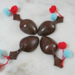 Puede incluir: Cuatro cucharas de madera marrón oscuro dispuestas en un patrón circular. Cada cuchara está adornada con un pompón rojo y azul claro, sujeto con un hilo rojo. Las cucharas están sobre una superficie blanca.