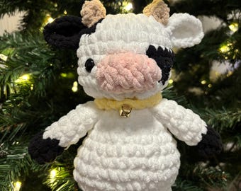 May include: A handmade crochet cow plushie with a white body, black spots, and black hooves. It has a pink muzzle, small brown horns, and a yellow collar with a small bell. The cow is standing in front of a green Christmas tree.