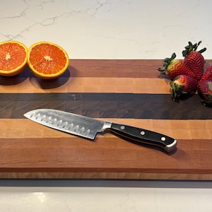 Può includere: Un tagliere di legno con un motivo a strisce in tonalità di marrone, rosso e giallo. Il tagliere ha un coltello e due metà di un'arancia sopra. Ci sono anche diverse fragole rosse sul tagliere.