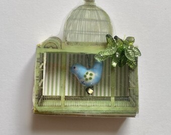 May include: A decorative sticker featuring a light green birdcage with a domed top and vertical striped background. Inside the cage is a small, light blue ceramic bird with green spots, and a cluster of green plastic leaves is attached to the side.