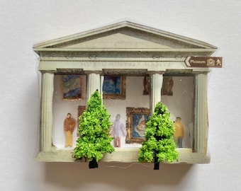 May include: A miniature museum building model featuring a classical facade with columns, two bright green model trees in the foreground, and interior walls decorated with small framed paintings and three tiny human figures.