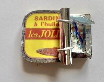 May include: A rectangular brooch shaped like an open sardine tin with a yellow label reading SARDINES à l'huile les JOLIES. The lid is partially peeled back to reveal a metallic interior with small, iridescent blue and silver fish figures.