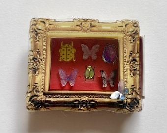 May include: A miniature gold-toned ornate frame containing a red velvet-like backing with six small insect and butterfly figures. The collection includes a yellow beetle, a purple beetle, a green beetle, and three translucent butterflies.