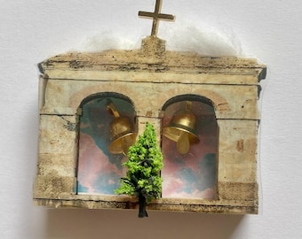 May include: A miniature beige stone-textured church facade ornament featuring two arched openings, each containing a small gold bell. A gold cross sits atop the structure, and a small green model tree is attached to the front center.