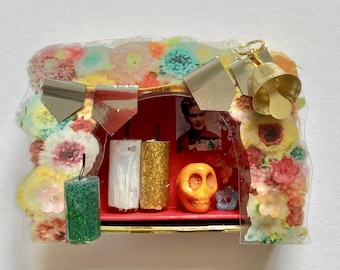 May include: A miniature diorama box featuring a floral border, a gold bell, and a red interior. Inside are three small cylindrical candles in green, white, and gold, an orange skull figurine, and a small portrait of Frida Kahlo.