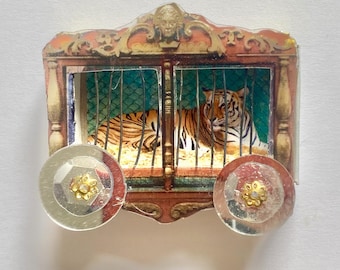 May include: A decorative brooch shaped like a circus wagon with a tiger inside a cage. The wagon is brown and gold with a detailed top crest and two circular, reflective silver wheels featuring small gold floral centers.