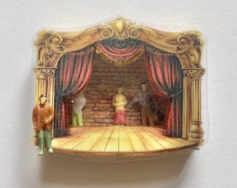 May include: A miniature theater stage pin featuring a gold-toned proscenium arch, red curtains, and a brick wall backdrop. The stage floor is light wood, and four small human figures are positioned on the stage and at the front edge.