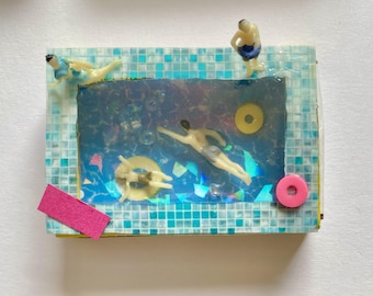 May include: A rectangular diorama featuring a miniature swimming pool with blue tiled borders. Inside the pool, three small figures are swimming or floating on yellow rings. A pink circular bead and a pink rectangular felt piece are attached.