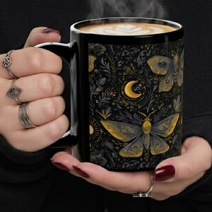 May include: Black coffee mug with a detailed moth and moon design in gold and gray. The mug is filled with coffee, and steam rises from the top. The mug is held in a person's hands.
