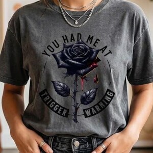 Può includere: T-shirt grigio scuro con una grafica di rosa nera e la scritta "YOU HAD ME AT TRIGGER WARNING". La maglietta ha un aspetto vintage ed è abbinata a jeans scuri.