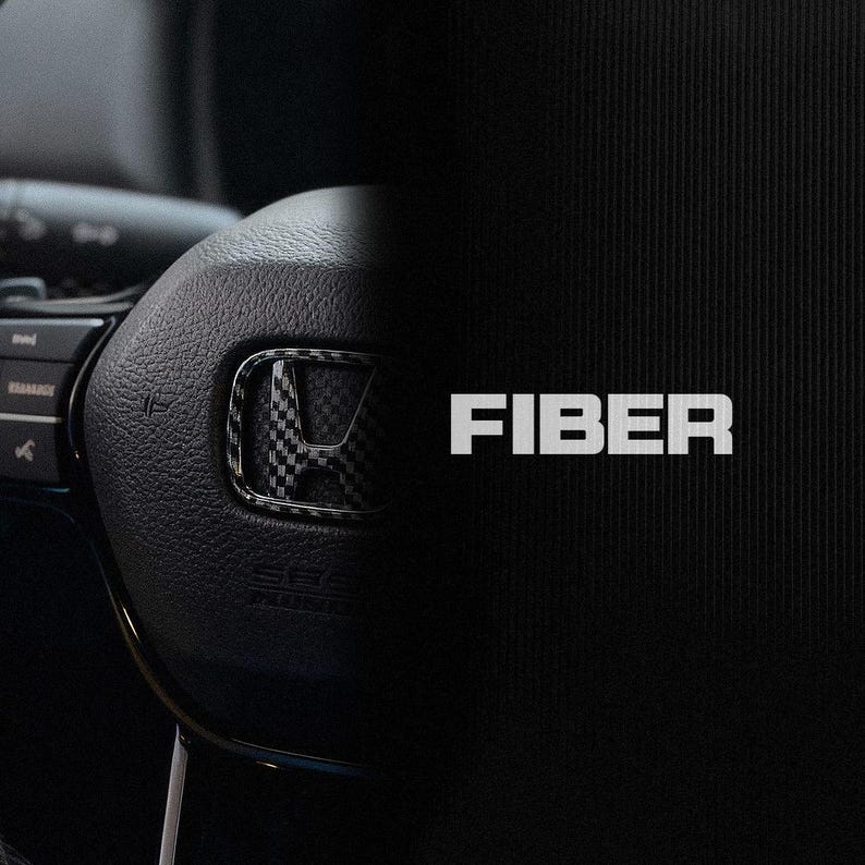 May include: A close-up of a black car steering wheel featuring a carbon fibre emblem. The word "FIBER" is displayed in white, bold letters on a black background. The steering wheel has buttons and controls.
