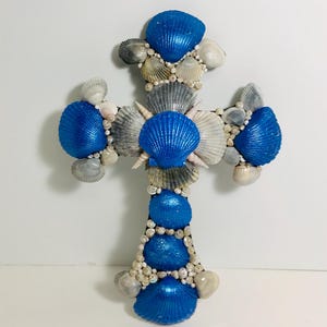 May include: A blue and white seashell cross made of various seashells. The cross is decorated with blue iridescent seashells.