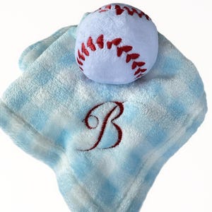 Puede incluir: Una manta de seguridad de felpa suave con un patrón de cuadros azul claro y blanco. En el centro hay una pequeña pelota de béisbol de felpa blanca con costuras rojas bordadas. La letra B está bordada con hilo rojo sobre la manta.