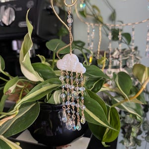 May include: A decorative gold-toned metal mobile featuring a white cloud with dangling crystal beads, resembling raindrops. The cloud is suspended from a curved wire, with additional crystal accents. The mobile is set against a backdrop of green plants.